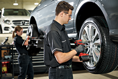 An image of two technicians performing a tire rotation and wheel alignment on a sedan. 
